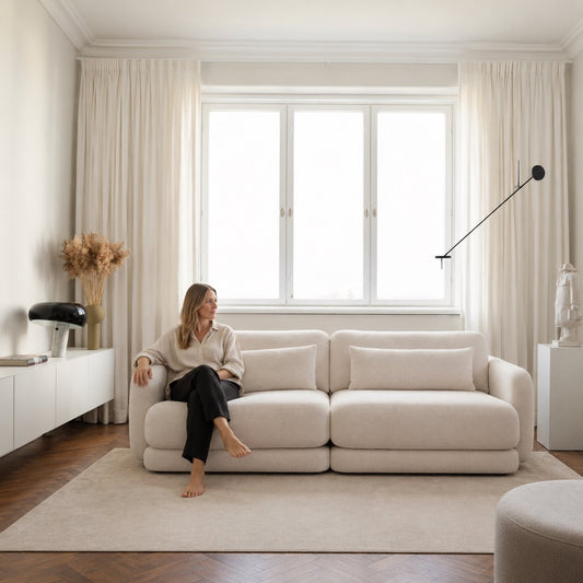 Junge Frau mit blonden Haaren sitzt entspannt auf Sofa BOHO 210, das auf einem hellen Teppichboden steht, Hersteller: Oot-Oot, Bild KI-generiert