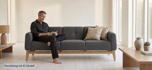 Dunkles 3-Sitzer Sofa Smooth aus Stoff im skandinavischen Design mit Holzbeinen, auf dem ein Mann sitzt. Hersteller: Tarmeko. Darstellung mit einem virtuellen KI-Modell.