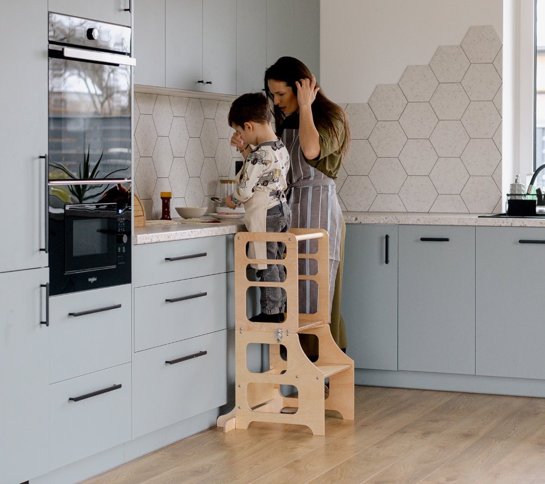 Eine Junge steht im multifunktionalen 2-in-1 Lernturm & Kindertisch in skandinavischem Design in der Farbe natur, Hersteller: Duck Woodworks