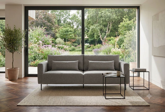 Schlafsofa FOLD mit grauem Bezug auf einem hellen Teppich vor einem großen Fenster mit Blick in den Garten, Hersteller: Oot-Oot, Bild: KI-generiert