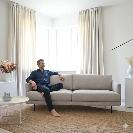 Sofa Floyd 220 cm auf dem ein Mann mit Bart lässig sitzt in einem hellen  Wohnzimmer vor einem großen Fenster, Hersteller: Oot-Oot, Bild: KI-generiert