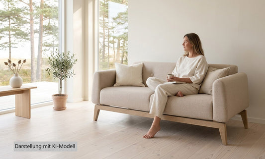 Modernes, helles Stoffsofa in einem lichtdurchfluteten Raum. Eine Frau in heller Leinenkleidung sitzt mit einer Kaffeetasse auf der Couch und blickt zum Fenster. Hinweis: Die abgebildete Person ist ein digital erzeugtes KI-Modell.