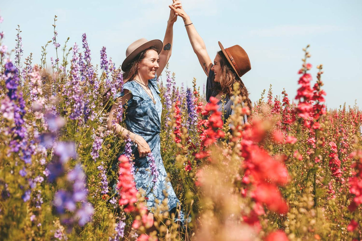 Zwei junge Frauen in bunten Kleidern und mit Hüten tanzend in einer Blumenwiese. Foto: Roxxie Blackham/Unsplash